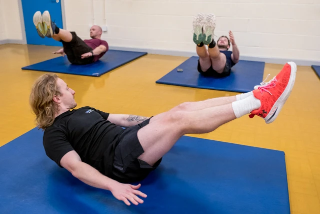 Total Abs (6) Class instructor shows Total Abs calss attendees how to perform abdominal exercises on yoga mats.