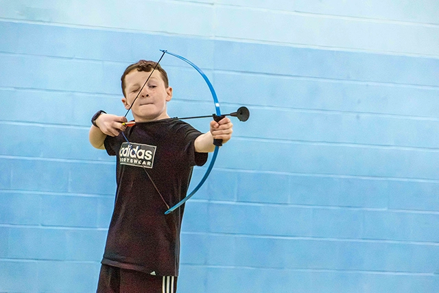 Archery A young boy in a black t-shirt draws back a blue bow, aiming forward. He is practicing archery in front of a light blue wall.