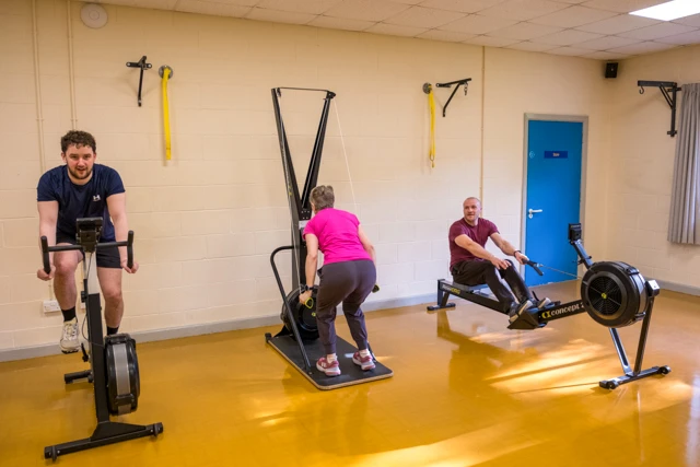 HIIT Three people are exercising during HIIT class using different machines. A man is on a stationary bike, a woman is on a ski machine, and another man is on a rowing machine.