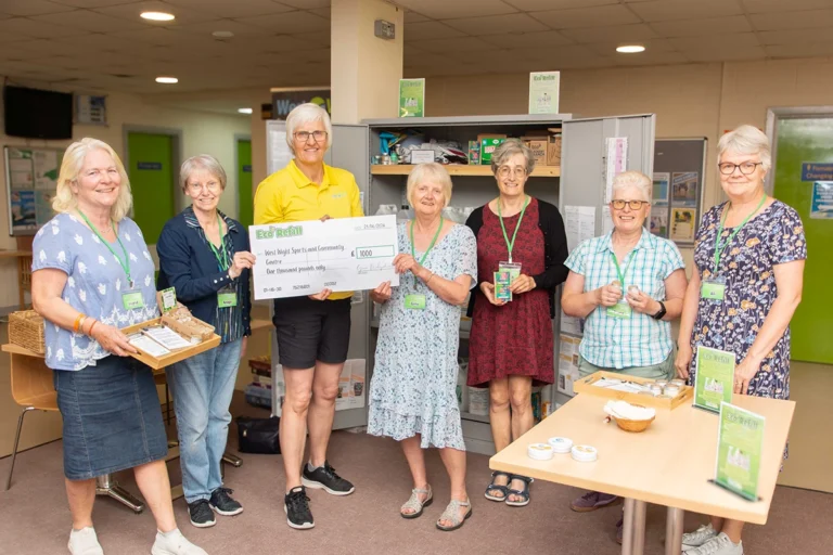 Eco Refill 29-06-2026 copy A group of female Eco Refill volunteers are posing for a photo, alongside centre manager, Clare Griffin. They are presenting a £1000 cheque. Several women are wearing lanyards with name tags. There are also items displayed on a table in the foreground, related to the eco-refill service.
