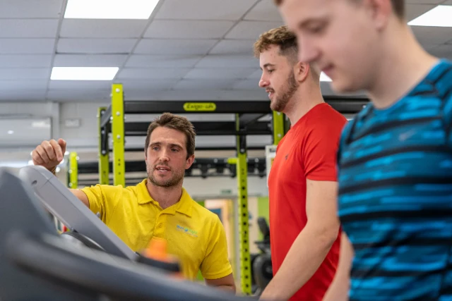 A fitness instructor in a yellow shirt points to a treadmill while two other men stand nearby in a gym.
